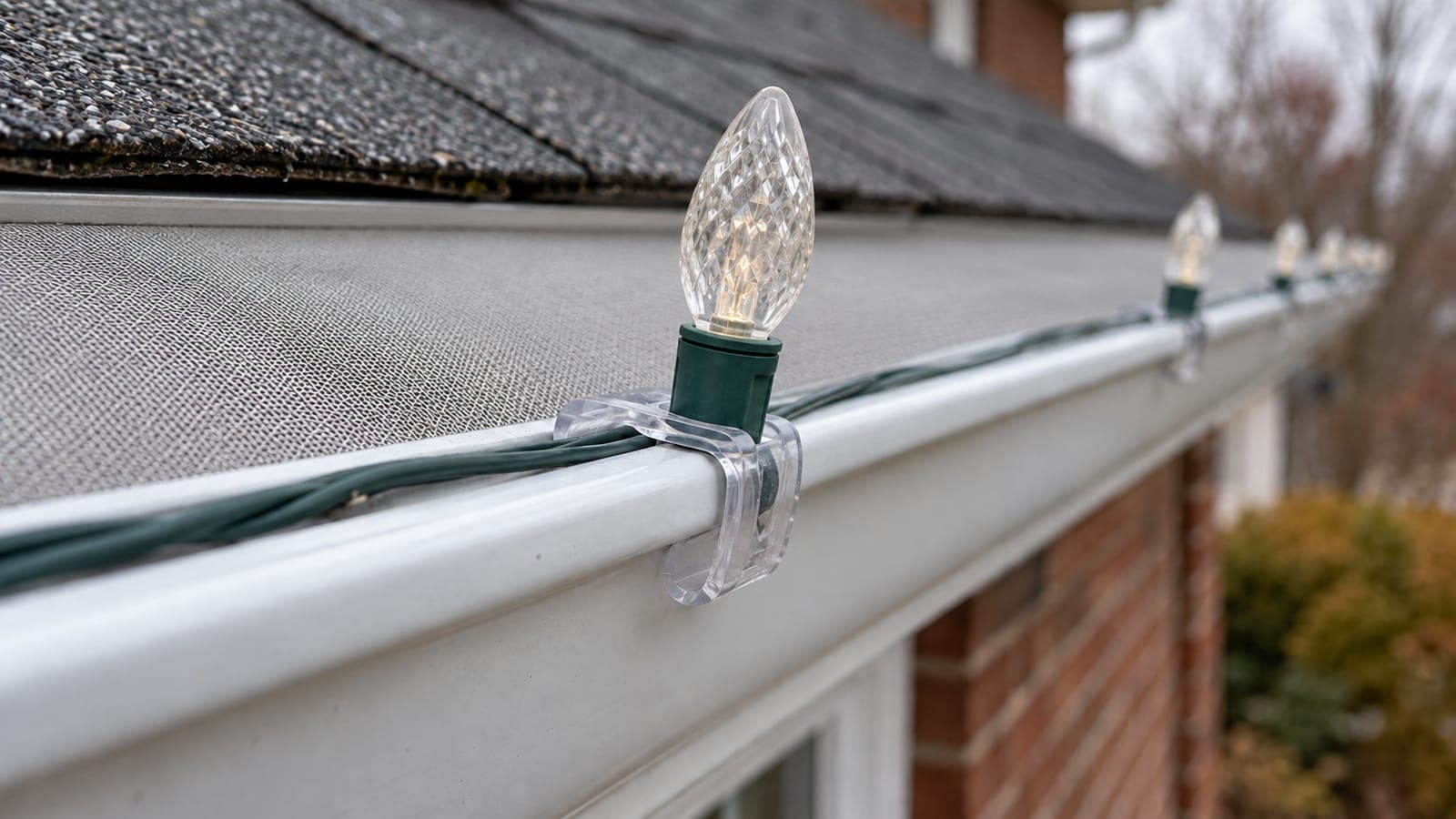 Clear plastic light clip hooked over the front edge of an eavestrough with a flat micro-mesh guard